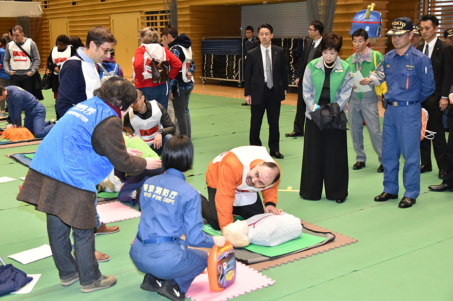 Governor Koike watches a foreign man practicing first aid on a mannequin.