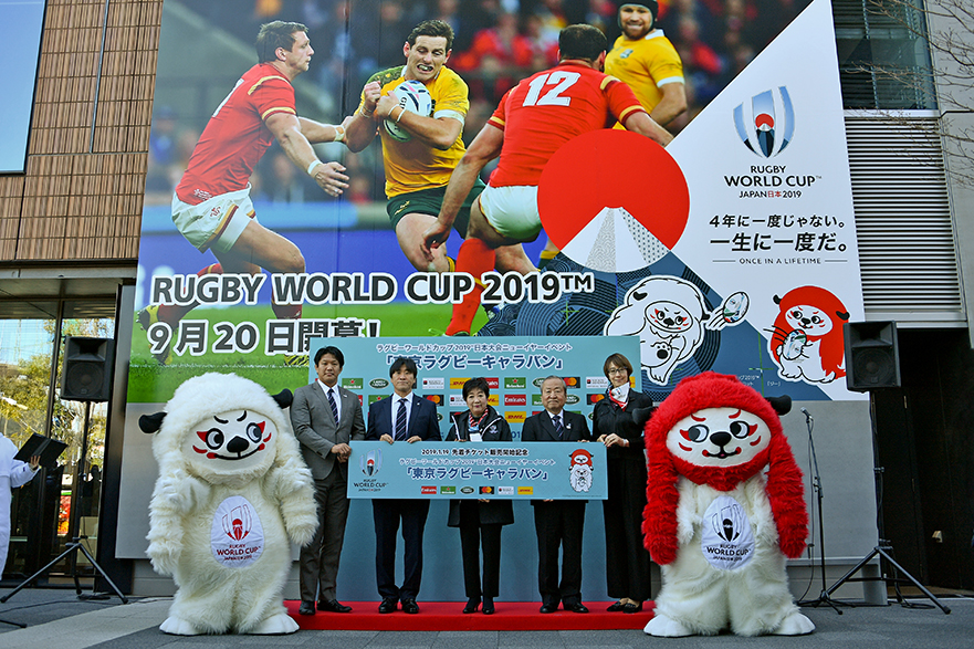 Governor Koike and others flanked by the Rugby World Cup mascots hold a sign.